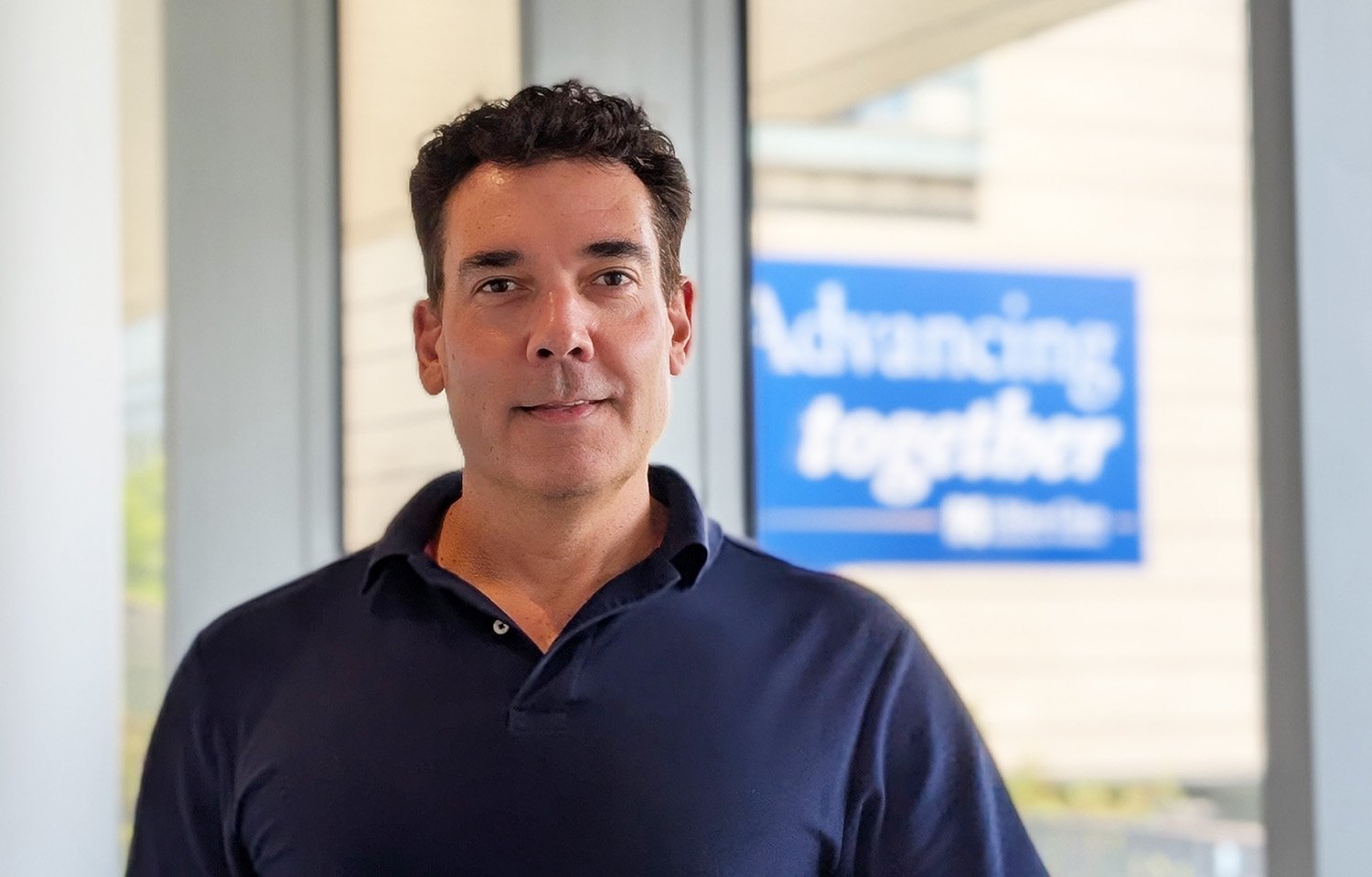 Man in blue shirt standing in front of a window with Advancing Together logo in background